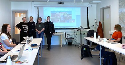 Mötesdeltagare runt ett hästskoformat bord, tittar mot fyra personer vid en whiteboard. 