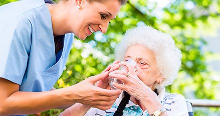 Hemtjänstpersonal hjälper äldre dam att dricka vatten i värmen.