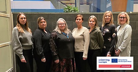 Gruppbild med sju kvinnor som är uppställda för fotografering inomhus.