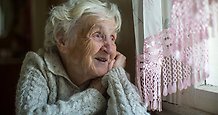 En äldre dam tittar ut genom fönstret med glädje i blicken. 